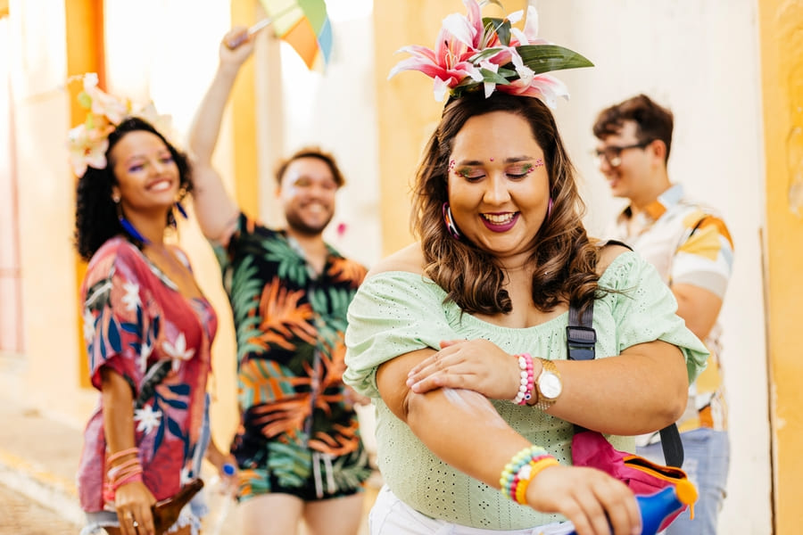 4 pessoas com adereços de carnaval: 1 mulher aplicando produto na pele do braço esquerdo e o restante dançando ao fundo
